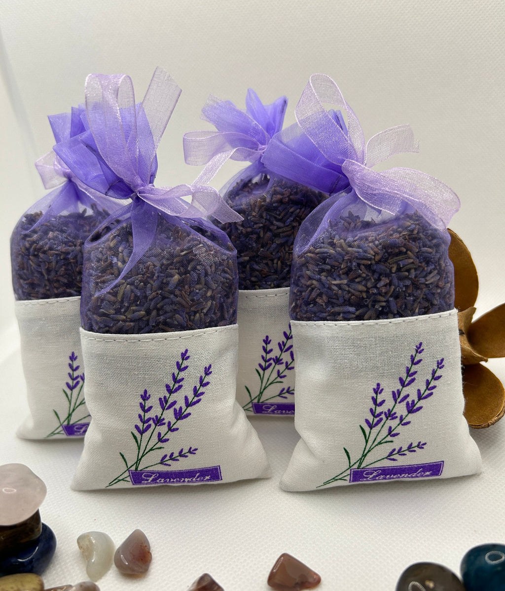 May include: Four white fabric lavender sachets with purple lavender flowers printed on them. Each sachet is tied with a purple ribbon and filled with dried lavender and lavender oil infused Epsom salt. The sachets are arranged in a row on a white surface. The text 'Lavender' is printed on the bottom of each sachet.