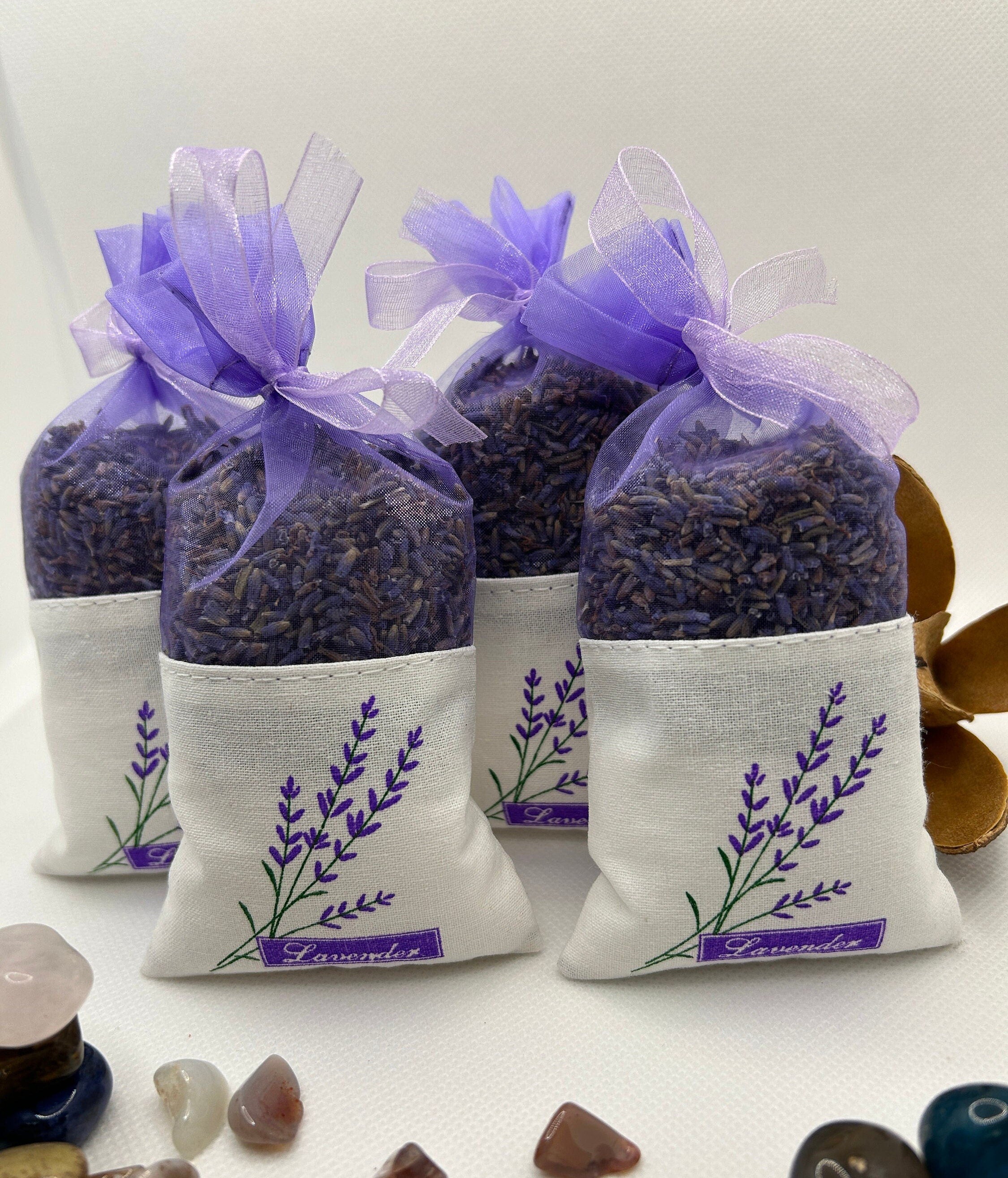 May include: Four white fabric lavender sachets with purple lavender flowers printed on them. Each sachet is tied with a purple ribbon and filled with dried lavender and lavender oil infused Epsom salt. The sachets are arranged in a row on a white surface. The text 'Lavender' is printed on the bottom of each sachet.
